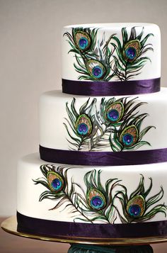 a three tiered cake with peacock feathers on the top and purple ribbon around the bottom