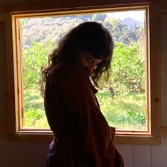 a woman standing in front of a window looking out at the trees and grass outside