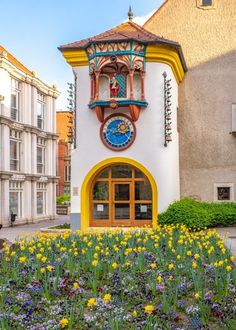 a building with a clock on the side of it's face surrounded by flowers