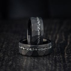 two wedding bands with diamonds on them sitting on a black surface, one is made out of wood and the other has silver inlays