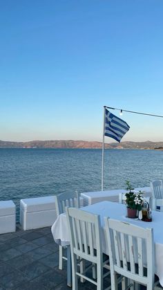 an outdoor dining area with tables and chairs near the water's edge, along with a greek flag flying in the background