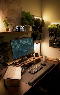 a desk with a computer, laptop and clock on it in front of some plants