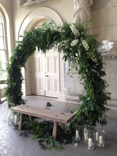 a wooden bench surrounded by greenery in front of a doorway with candles on the floor