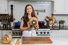 a woman holding a loaf of bread in front of a kitchen counter with white cabinets