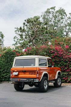 an orange and white jeep parked in front of some bushes with pink flowers behind it