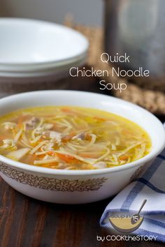 a bowl of chicken noodle soup on top of a wooden table next to plates