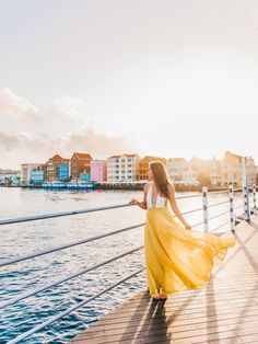 a woman in a yellow dress is walking on a pier near the water and buildings