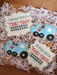 three decorated cookies in a box with white shredded paper on the bottom and one cookie shaped like a tractor