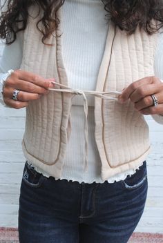 a woman is holding something in her hands while wearing a sweater vest and blue jeans