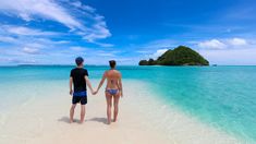 two people holding hands while standing on the beach in front of an island with clear blue water