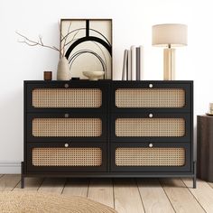 a black dresser with wicker drawers in a white room next to a lamp and pictures on the wall