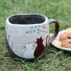 two mugs with cats on them sitting in the grass next to cookies and muffins