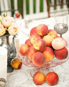 peaches in a glass bowl on a table with candles and flowers behind it,