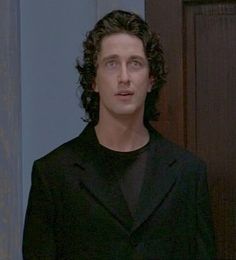 a man with curly hair wearing a black suit and standing in front of a door