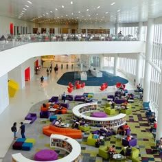 people are sitting and standing in the middle of a large room with colorful chairs, tables, and bookshelves