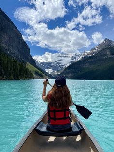 a woman in a red life jacket paddling a canoe on a lake with mountains in the background