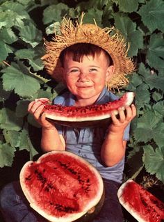 a young boy holding two slices of watermelon in front of his face and wearing a straw hat