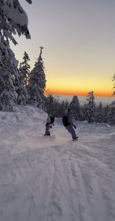 two snowboarders are going down a snowy hill at sunset with trees in the background