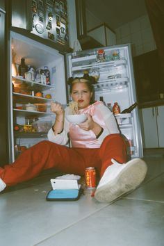 a woman sitting in front of an open refrigerator eating food and drinking from a cup