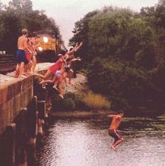 several people jumping into the water from a bridge over a body of water with a train in the background