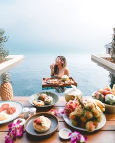 a woman sitting at a table with food and drinks in front of her, overlooking the ocean