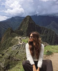 a woman sitting on top of a mountain next to a lush green hillside covered in clouds