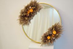 a round mirror with dried flowers in front of it on a white shelf next to a wall