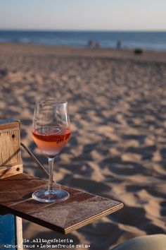 a glass of wine sitting on top of a wooden table next to the ocean with people in the background