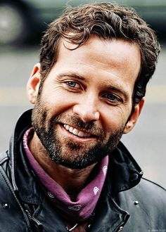 a close up of a person wearing a leather jacket and smiling at the camera with a car in the background