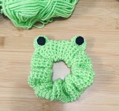 a crocheted green frog ring sits next to a ball of yarn on a wooden surface