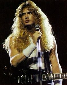 a man with long blonde hair holding a guitar and singing into a microphone on stage