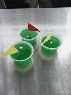 three plastic cups with green liquid and yellow paper flags sticking out of the top, on a metal surface