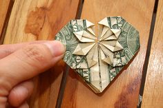 a hand holding a folded dollar bill in front of a heart shaped origami