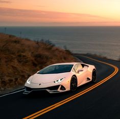 a white sports car driving down the road next to the ocean in front of a sunset