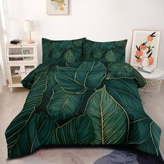 a bed with green leaves on it next to a white table and pictures hanging on the wall