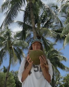 a woman is holding a coconut in front of her face while standing under palm trees