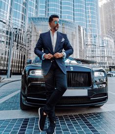 a man sitting on the hood of a black car in front of tall skyscrapers