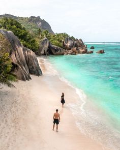 two people are walking along the beach in front of some rocks and clear blue water