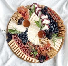 a platter filled with cheese, fruit and crackers on top of a white blanket