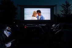 a couple kissing in front of a movie screen at night with cars parked around them