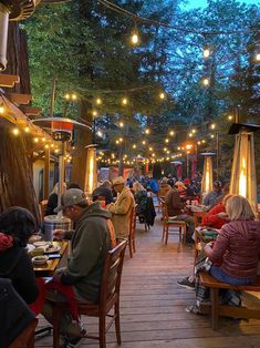 many people are sitting at tables and eating food in an outdoor restaurant with lights strung from the trees