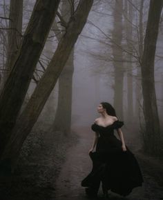 a woman in a black dress is walking through the woods on a foggy day