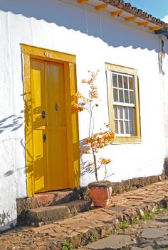 a white house with yellow door and window on cobblestone walkway next to potted plant