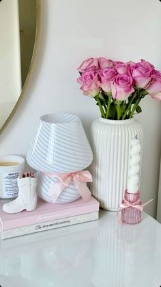 a white vase with pink roses on top of a table next to books and a mirror