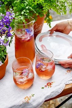 a table topped with two glasses filled with liquid and flowers next to potted plants