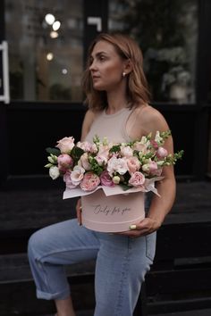 a woman sitting on a bench holding a pink box filled with flowers and greenery