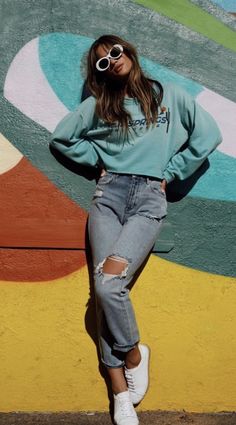 a woman standing in front of a colorful wall with her hands on her hips wearing ripped jeans and white sneakers