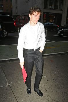 a young man in white shirt and black pants holding a red bag while standing on the sidewalk