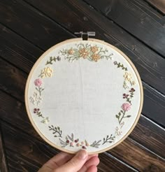 a hand is holding up a cross stitched embroidery hoop with flowers on it, which has been placed in front of a wooden wall