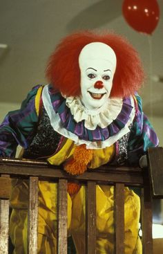 a clown with red hair sitting on top of a wooden bench